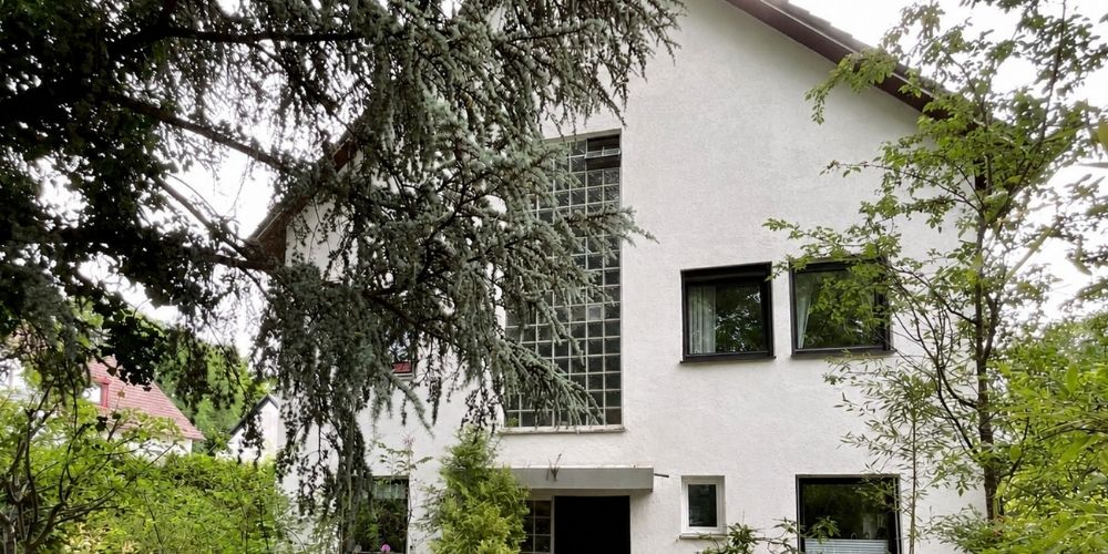 Weiße Außenfassade eines Einfamilienhauses mit großen Fenstern, Glasbausteinen und grüner Bepflanzung im Gartenbereich