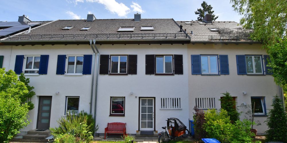 Weiße Reihenhausfassade mit farbigen Fensterläden, Dachflächenfenstern, Gartenbereich und begrüntem Außenbereich einer Münchner Immobilie