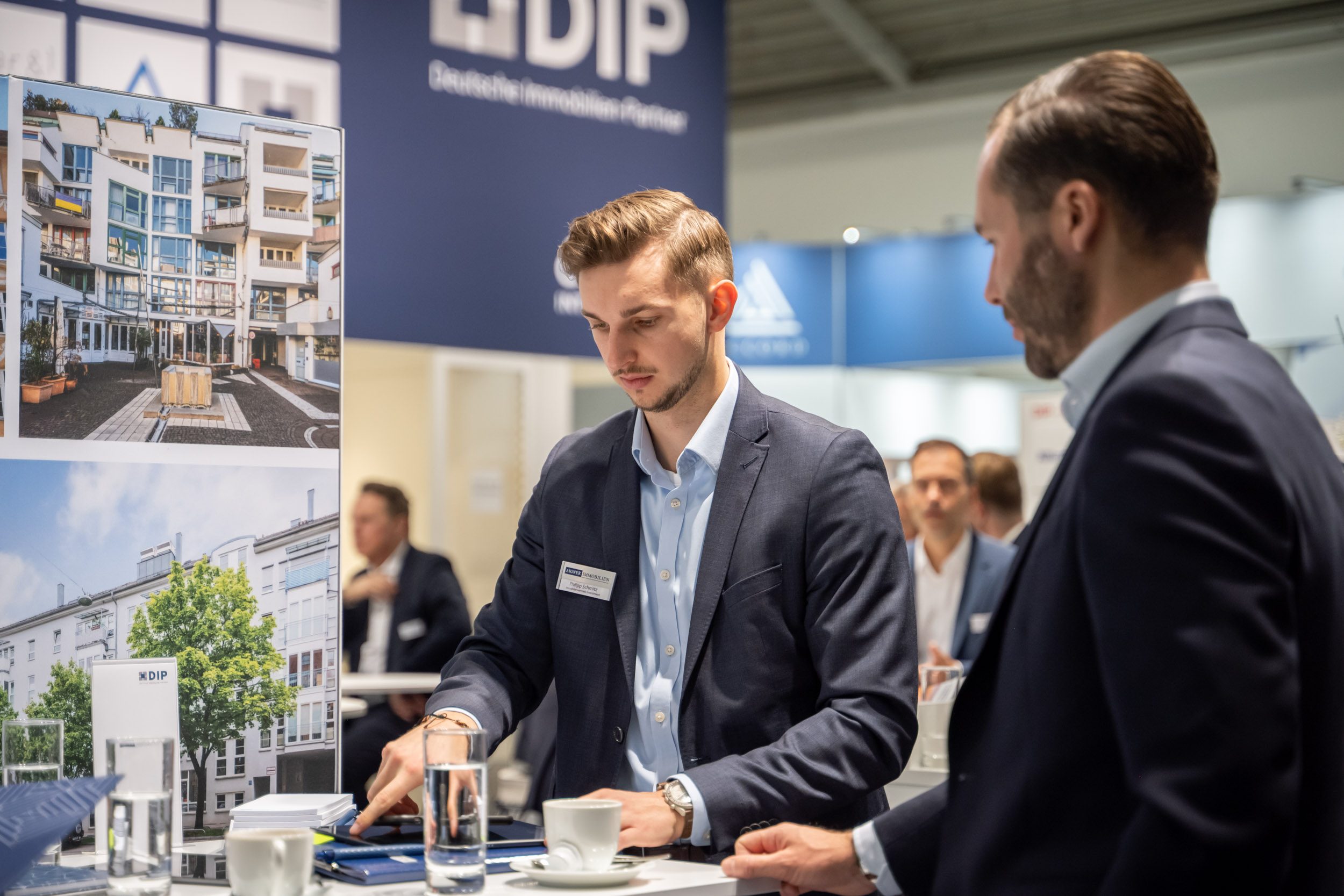 Investment-Objekte am Tablet präsentiert Berater tippt am Stehtisch auf ein Tablet, neben ihm ein Gesprächspartner, links Plakate mit Wohn- und Geschäftshäusern