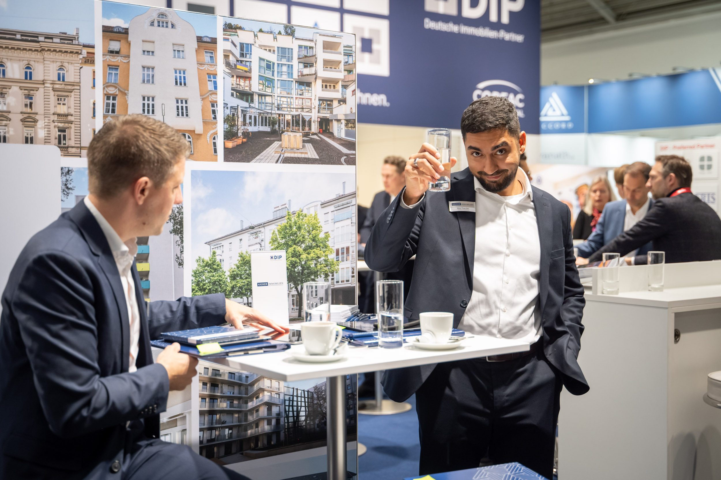 Prost mit Wasser am Besprechungstisch Mitarbeiter hebt am Tisch sein Wasserglas, gegenüber sitzt ein Gast, dahinter Fotos von Altbau- und Neubaufassaden