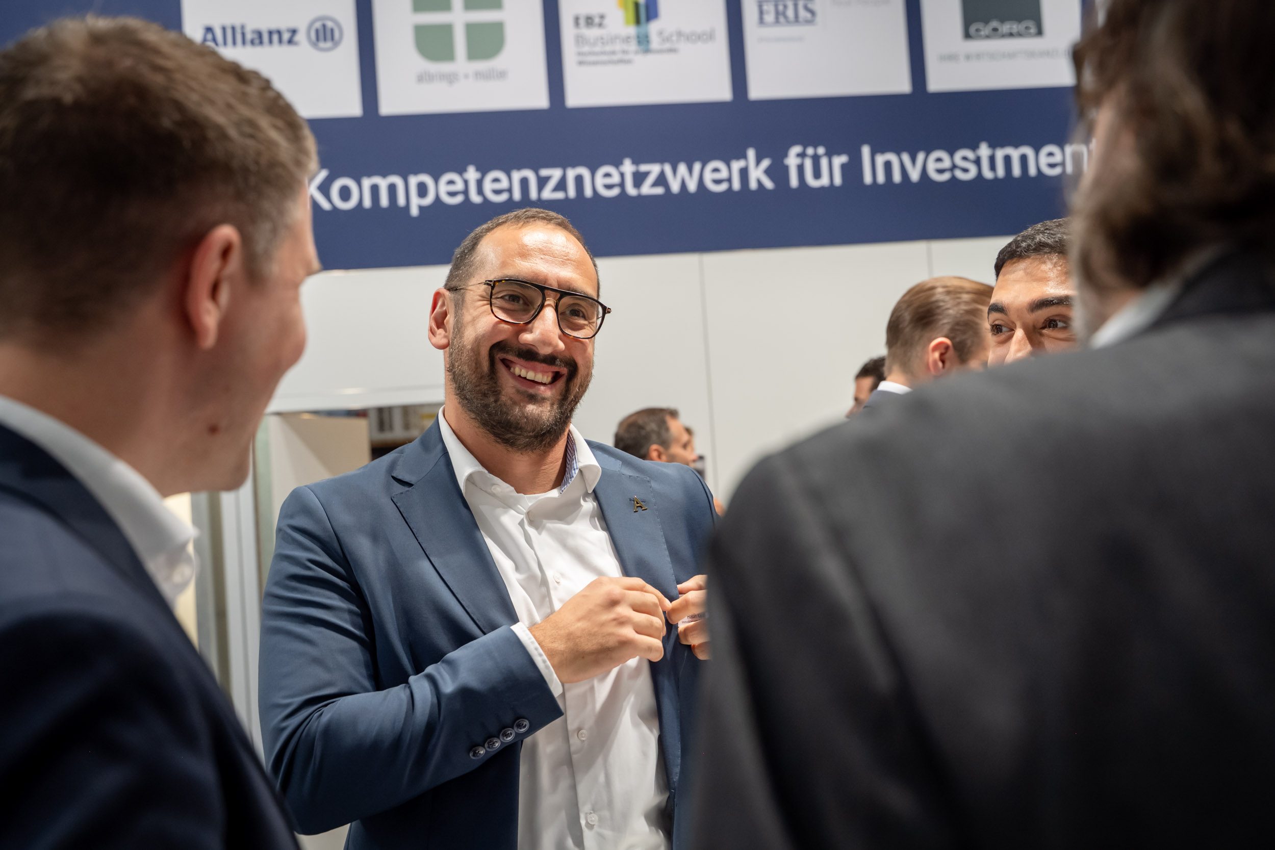 Gute Stimmung unter dem Netzwerkbanner Lachender Mann mit Brille und Bart steckt sein Namensschild an, darüber Banner „Kompetenznetzwerk für Investment“