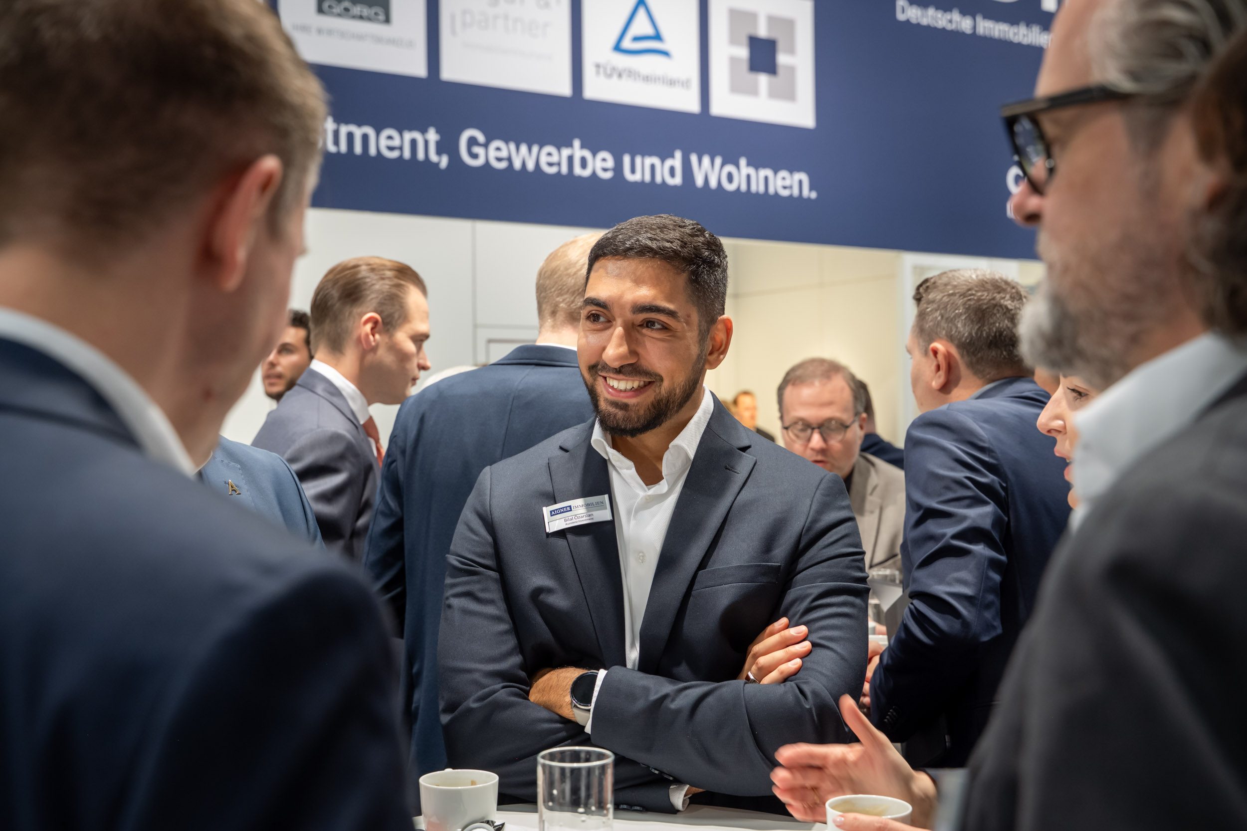 Lockeres Gespräch am Stehtisch Investment-Assistent mit verschränkten Armen lächelt im Gespräch am Stehtisch, im Hintergrund dichtes Messetreiben