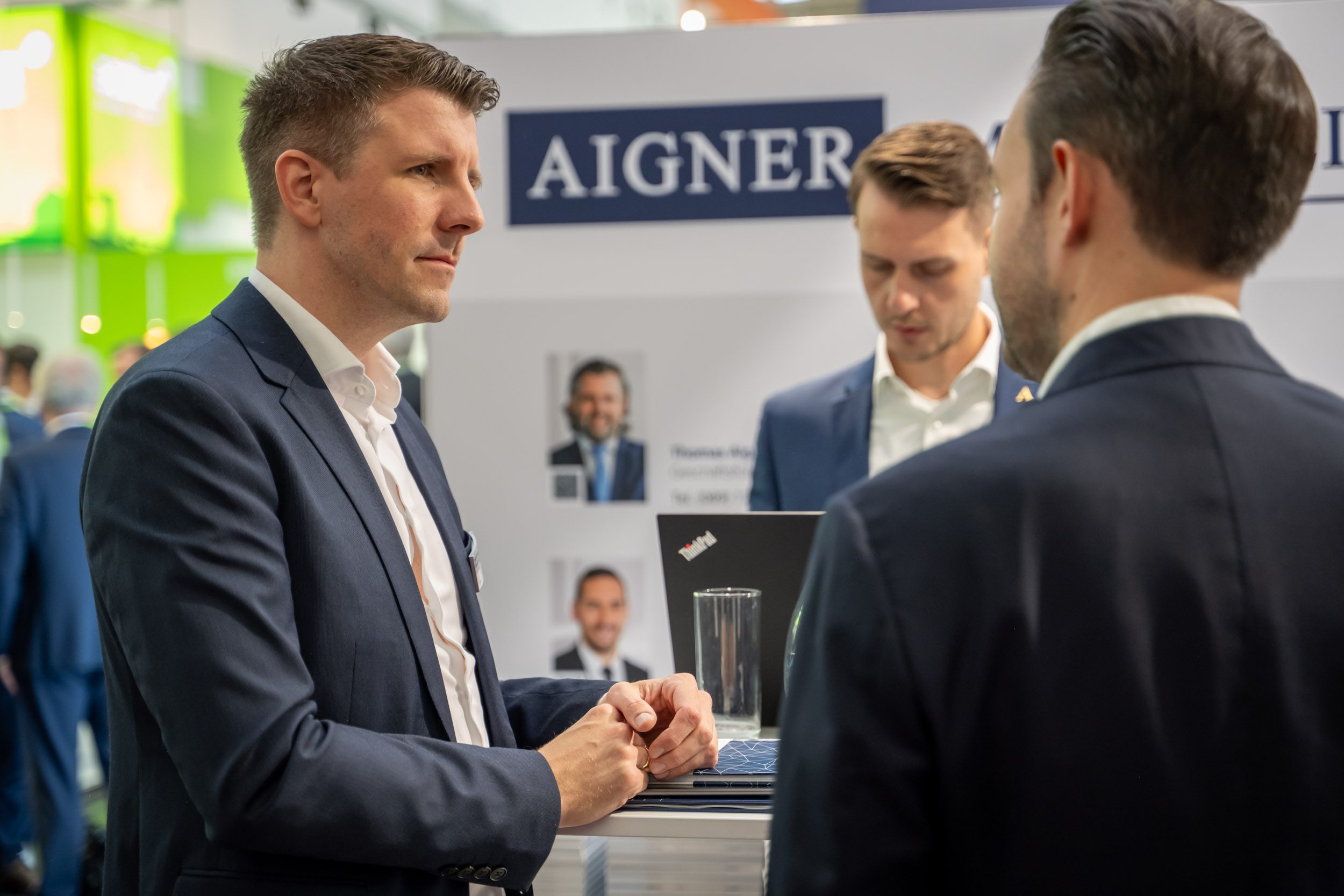 Zuhören vor der Teamwand Mann im dunkelblauen Anzug hört konzentriert zu, dahinter Aigner-Standwand mit Porträtfotos von Ansprechpartnern