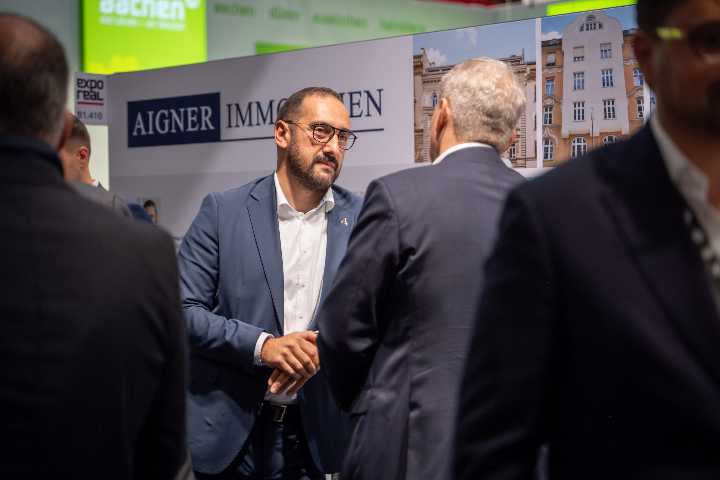 Handschlag am Stand B1.410 der EXPO REAL Handschlag zwischen Mann mit Brille und grauhaarigem Besucher vor Aigner-Immobilien-Wand, links Schild „expo real B1.410“