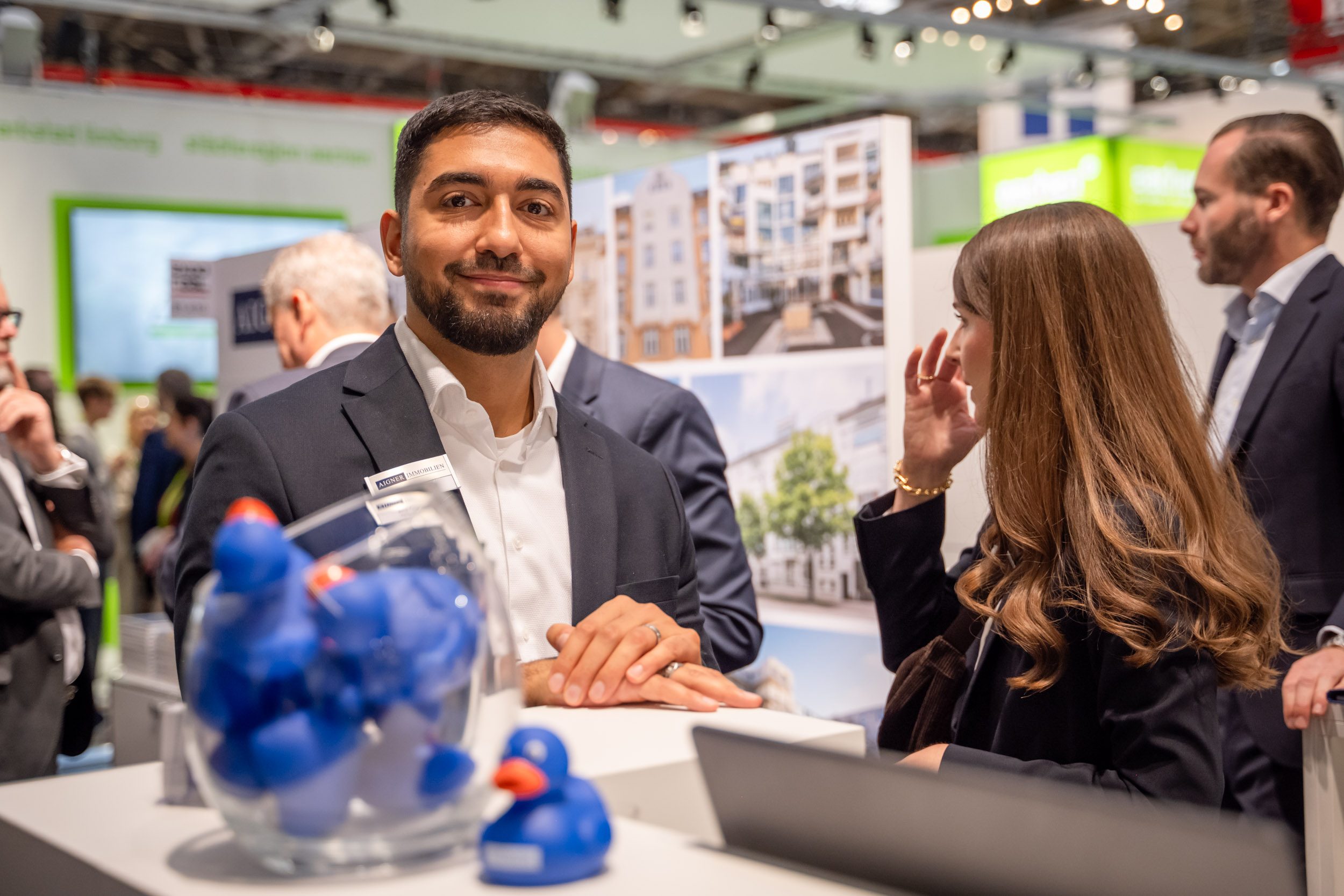Messestand mit blauen Quietscheenten Lächelnder Mitarbeiter am Messetresen, davor blaue Quietscheenten in Glasschale, im Hintergrund Fotos von Wohngebäuden