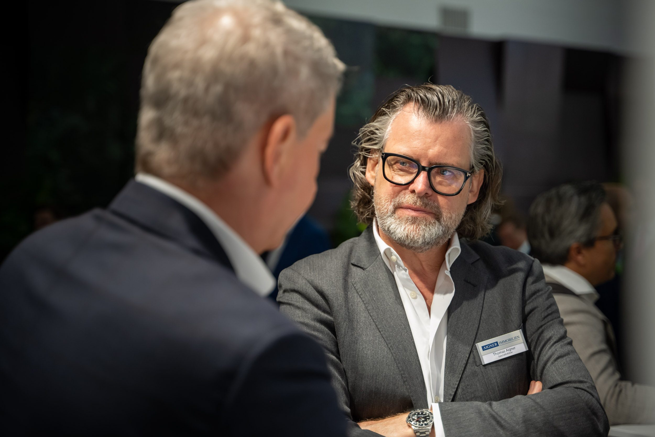 Geschäftsführer hört aufmerksam zu Geschäftsführer von Aigner Immobilien mit Brille und verschränkten Armen hört einem Gesprächspartner aufmerksam zu