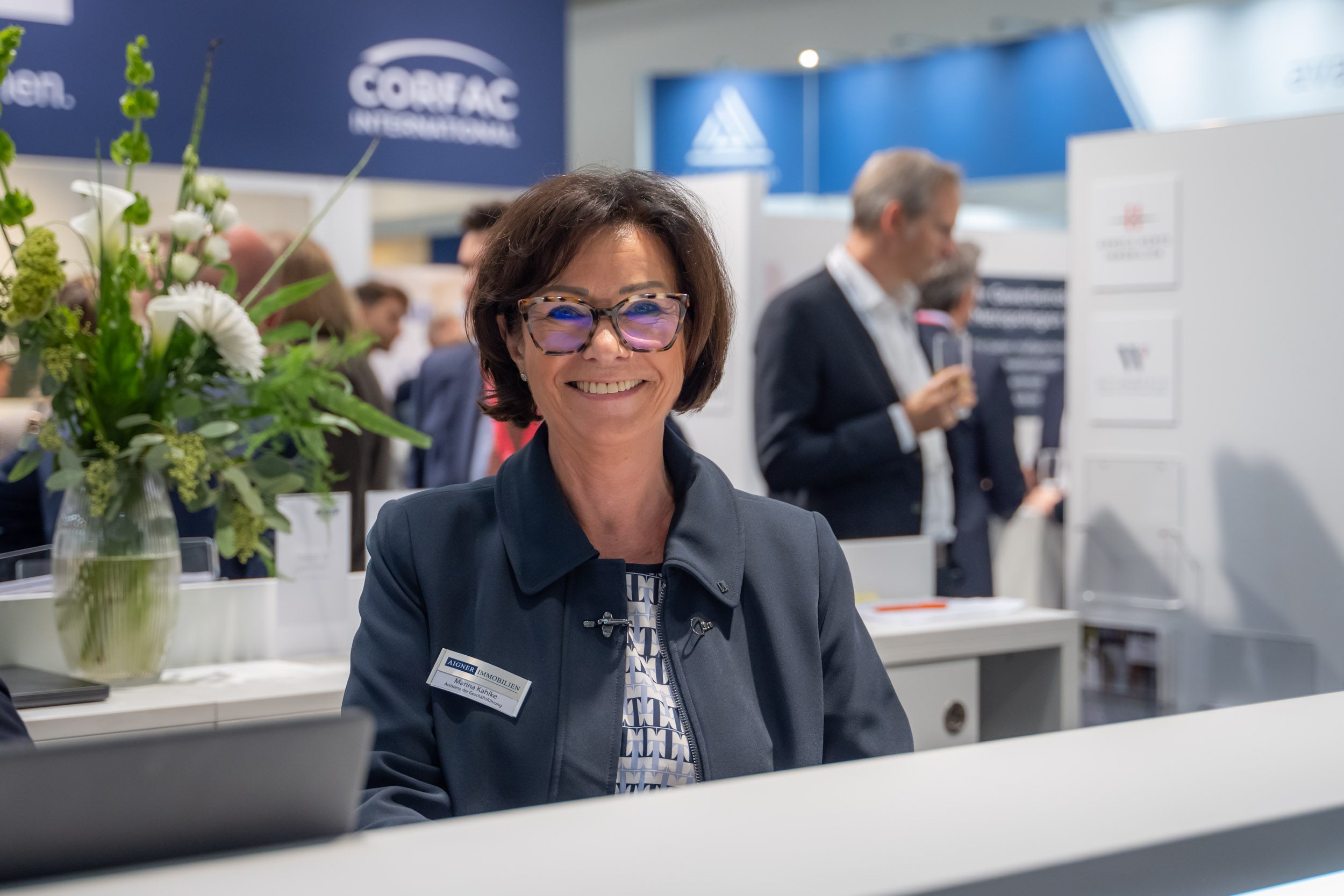 Freundlicher Empfang am Messetresen Lächelnde Assistentin der Geschäftsführung mit Brille am Empfangstresen des Messestands, daneben Blumenstrauß
