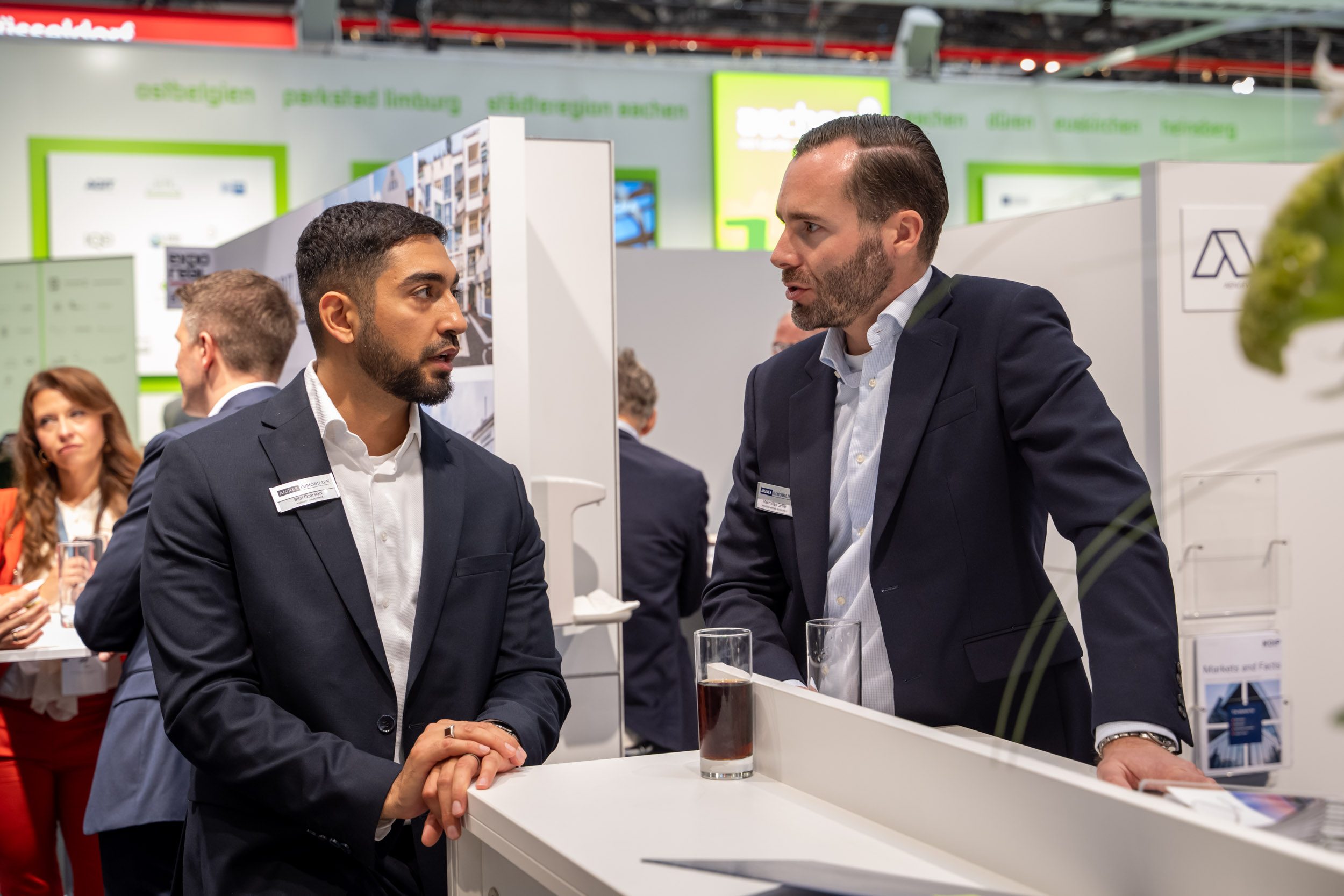 Austausch zweier Kollegen auf der EXPO REAL Zwei Mitarbeiter mit Aigner-Namensschildern im Gespräch am Stehtresen, im Hintergrund weitere Messestände