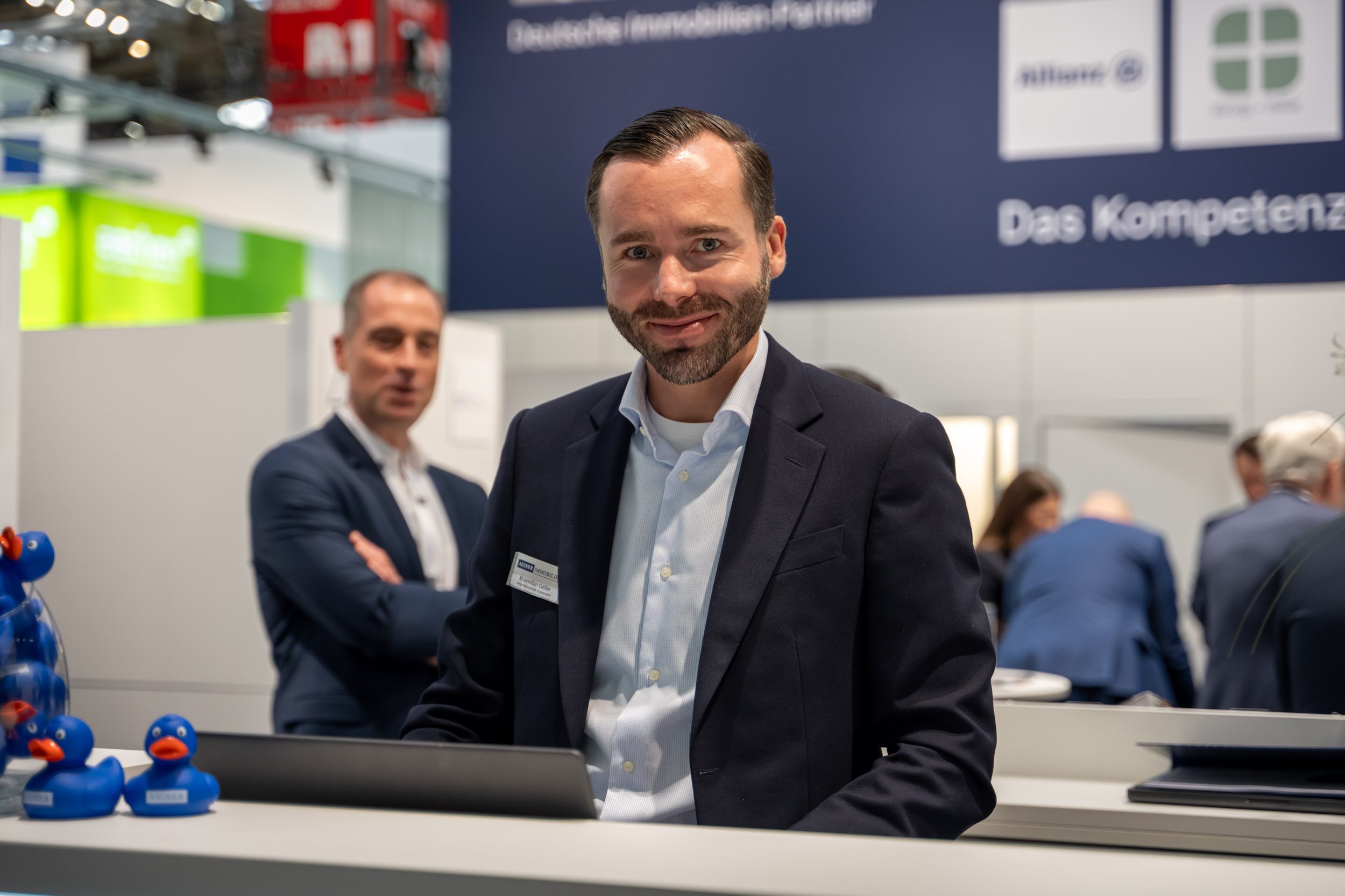Ansprechpartner hinter dem Messetresen Mitarbeiter mit Bart lächelt hinter dem Messetresen in die Kamera, vorne blaue Quietscheenten mit Aigner-Aufdruck