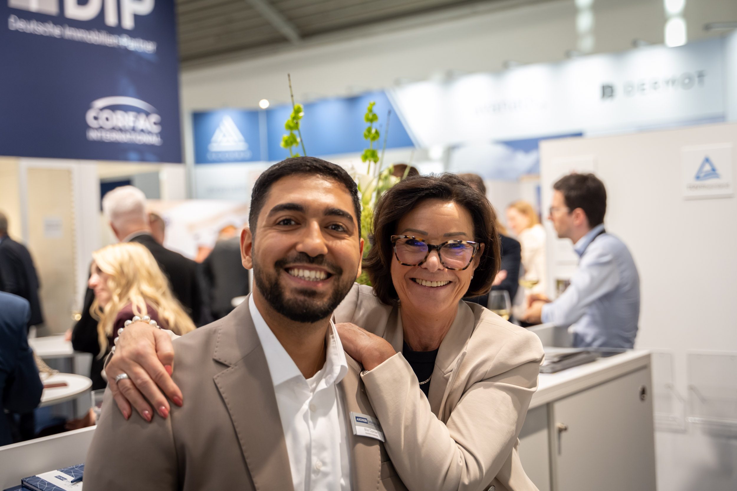 Teamspirit am Messestand Mitarbeiter und Kollegin in beigen Blazern lachen gemeinsam in die Kamera, sie legt ihm die Hände auf die Schulter