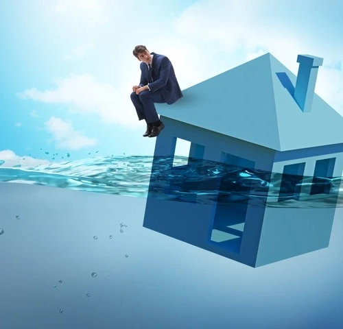 Symbolbild von einem Mann, der auf einem Haus sitzt, was im Wasser schwimmt und die Privatinsolvenz darstellt