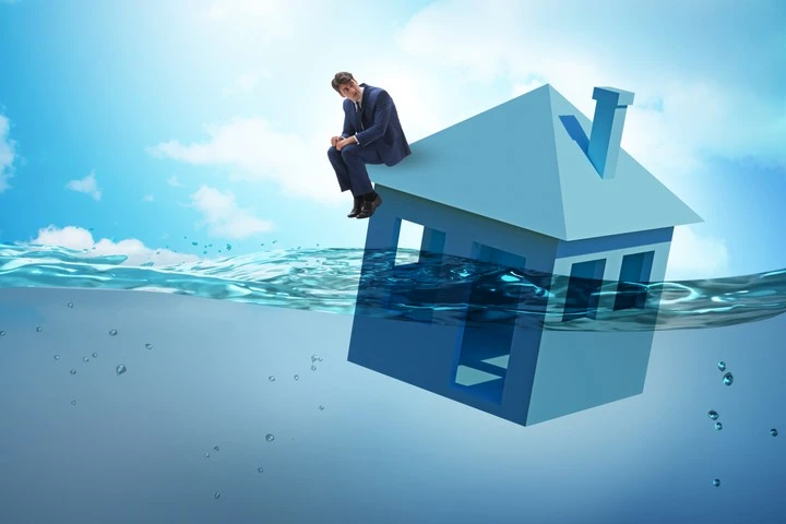 Symbolbild von einem Mann, der auf einem Haus sitzt, was im Wasser schwimmt und die Privatinsolvenz darstellt