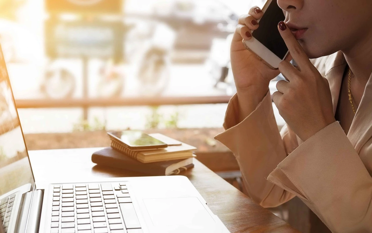Secret Sale - Frau sitzt vor dem PC und telefoniert mit vorgehaltenem Finger