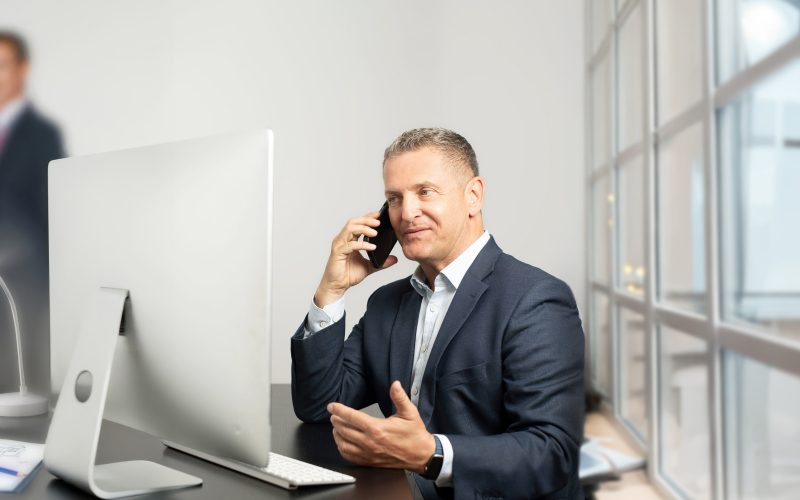Ralf Krebs beim Arbeiten im Büro vor einem PC, mit einem Handy in der Hand.