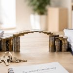 Eine Brücke aus aufgestapelten Euro-Münzen verbindet zwei Aktenordner mit Immobilien-Unterlagen auf einem Schreibtisch. Eine solide Zwischenfinanzierung schlägt die finanzielle Brücke beim Immobilienwechsel.