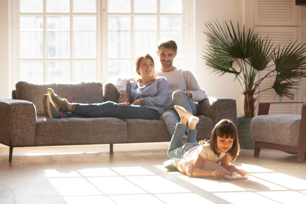 Familie sitzt gemütlich im Wohnzimmer auf der Couch, Mädchen spielt auf dem Boden